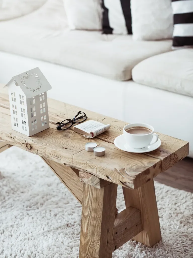 Table basse en bois avec une petite maison décorative, une tasse de café et des lunettes, devant un canapé clair
